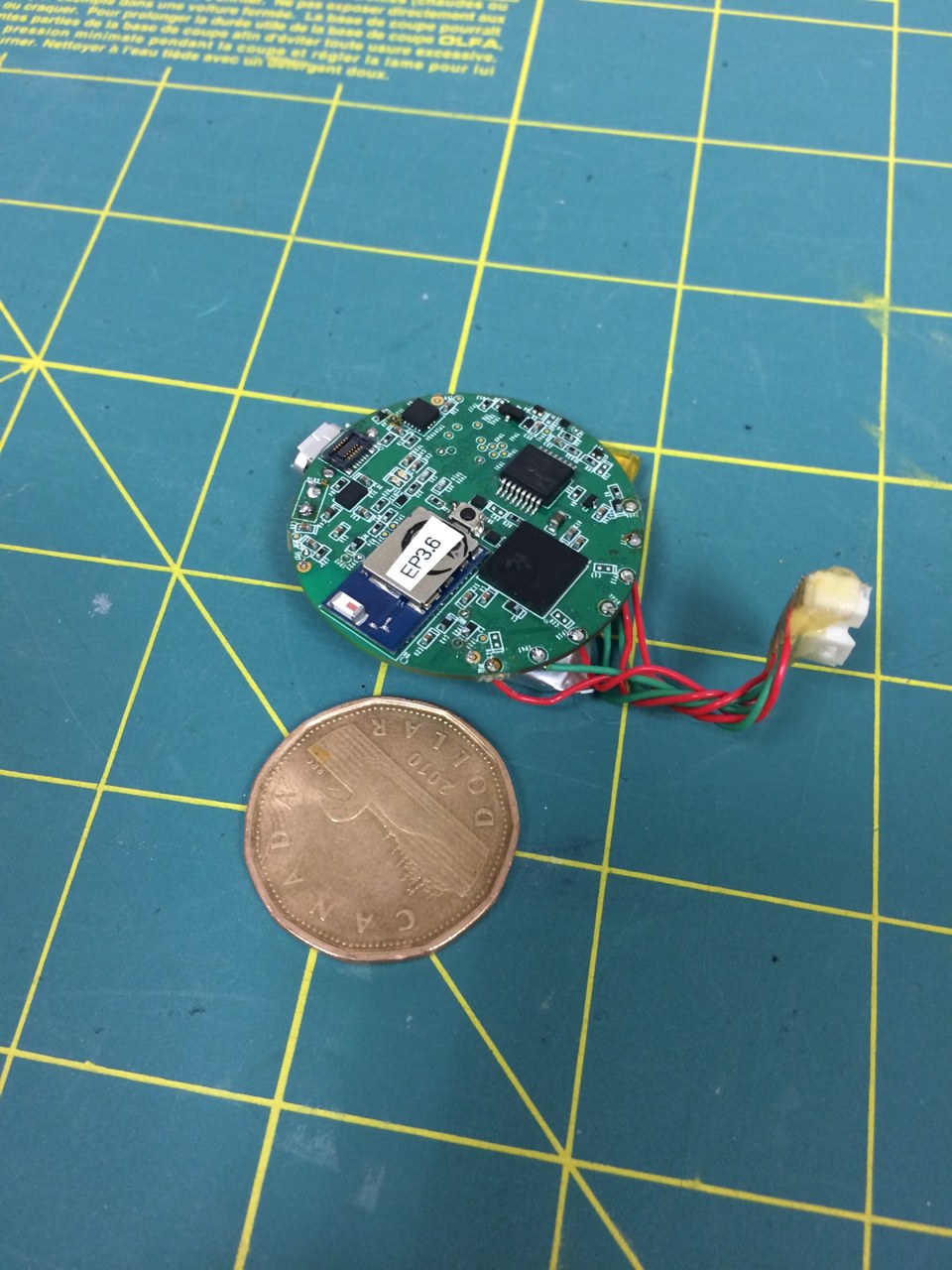 One of the earlier prototypes: a populated circular circuit board with attached wires and a coin providing a sense of scale.