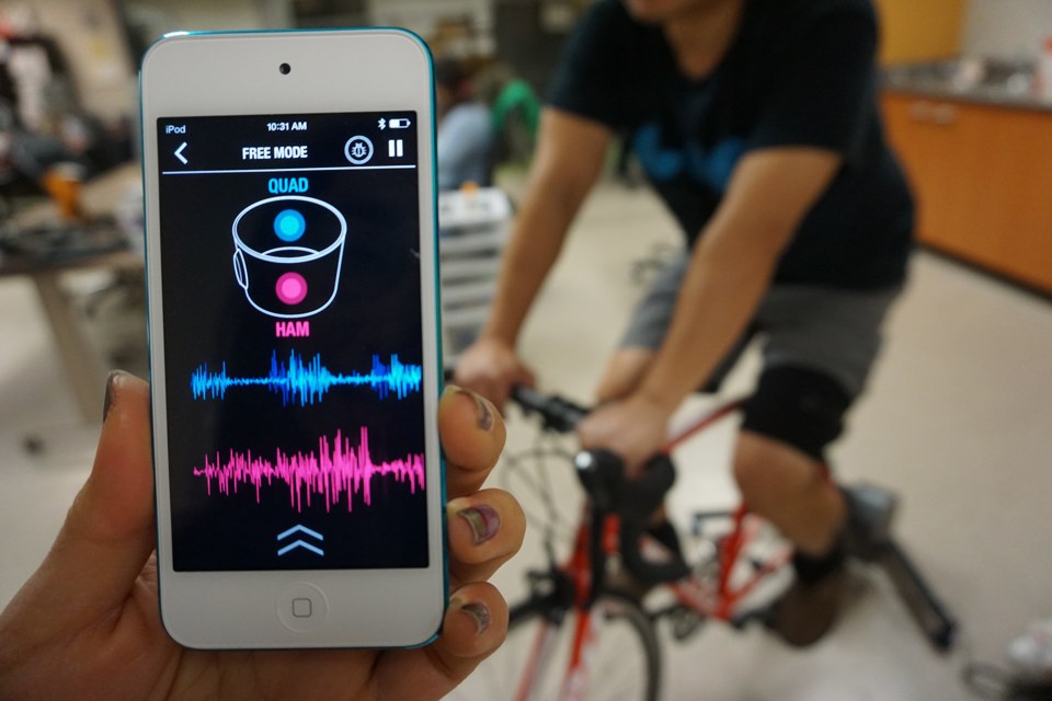 The mobile display shows separate quadriceps and hamstring traces while a rider pedals a bicycle.