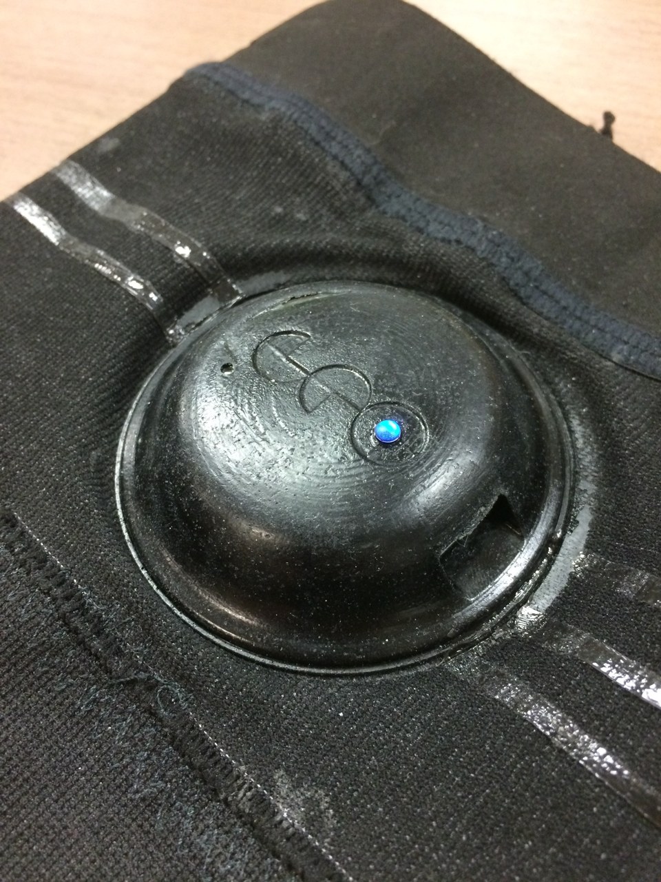 A black circular enclosure mounted in the textile band, with its blue indicator illuminated. This garment-integration approach was the subject of a patent application, filed but not pursued to issuance.