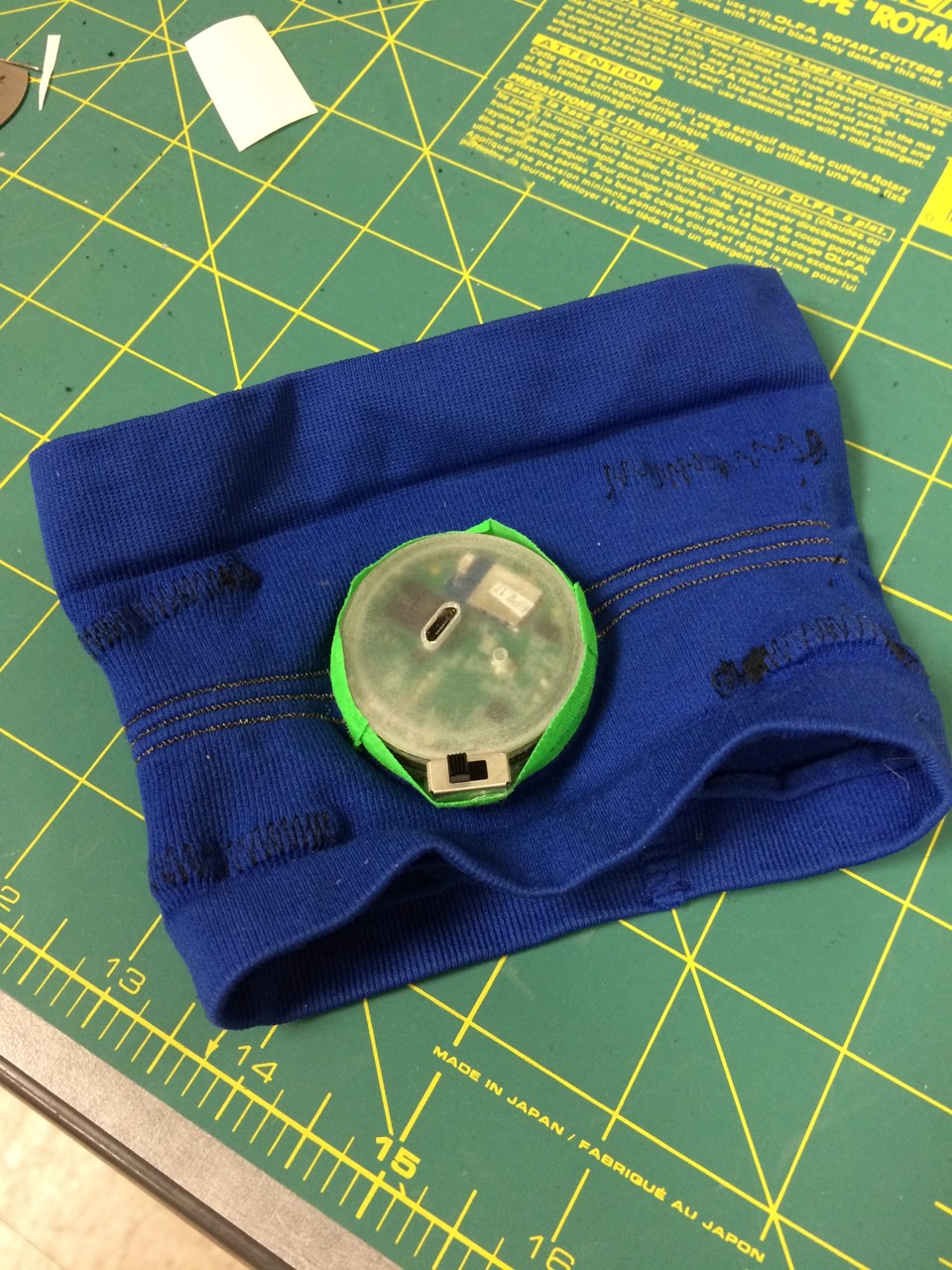 A transparent electronics module mounted on a blue textile band. Conductive textile connections link the module to conductive textile electrodes.
